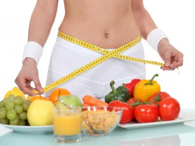 Mulher utilizando fita métrica em volta do corpo, junto a uma mesa com vegetais e frutas