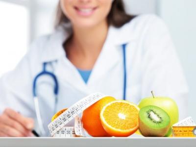 Imagem mostra uma nutricionista sentada em uma mesa que tem frutas e uma fita métrica em cima