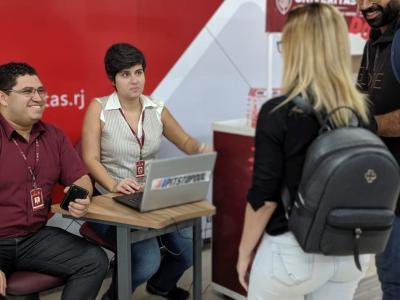 Imagem mostra uma mulher em pé e um homem e outra mulher sentados em frente a um computador